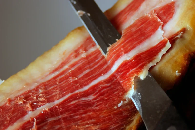 A knife carving a thin slice from jamón ibérico, showing the marbled red meat and white fat