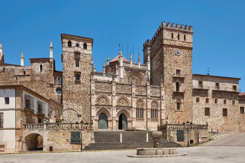 The ornate Gothic and Mudéjar facade of the Royal Monastery of Guadalupe, flanked by stone towers, seen across the empty square